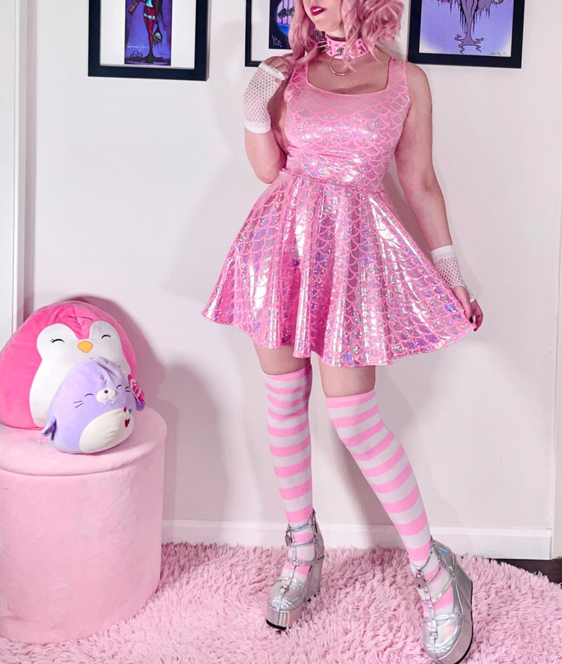 May include: A pink iridescent dress with a mermaid scale pattern. The dress has a scoop neck and is sleeveless. The model is wearing white striped knee-high socks and silver platform shoes.