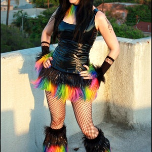 May include: A woman wearing a black leather top with a rainbow fur trim skirt and rainbow leg warmers. She is standing on a concrete surface with a city skyline in the background.