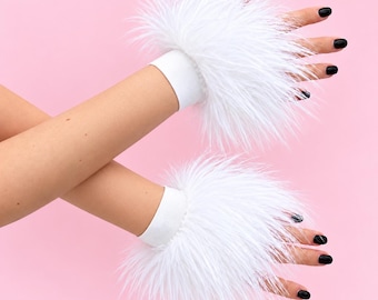 May include: A pair of white faux fur wrist cuffs featuring a soft, long-pile texture and a plain white elastic band base. The cuffs are positioned around the wrists, showcasing the fluffy white material against a solid light pink background.