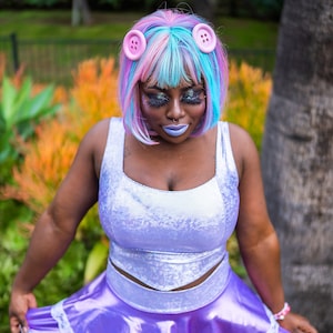 May include: A woman with pink and blue hair wearing a light purple, sparkly, sleeveless dress with a white bodice and a ruffled skirt. She is wearing pink buttons in her hair.
