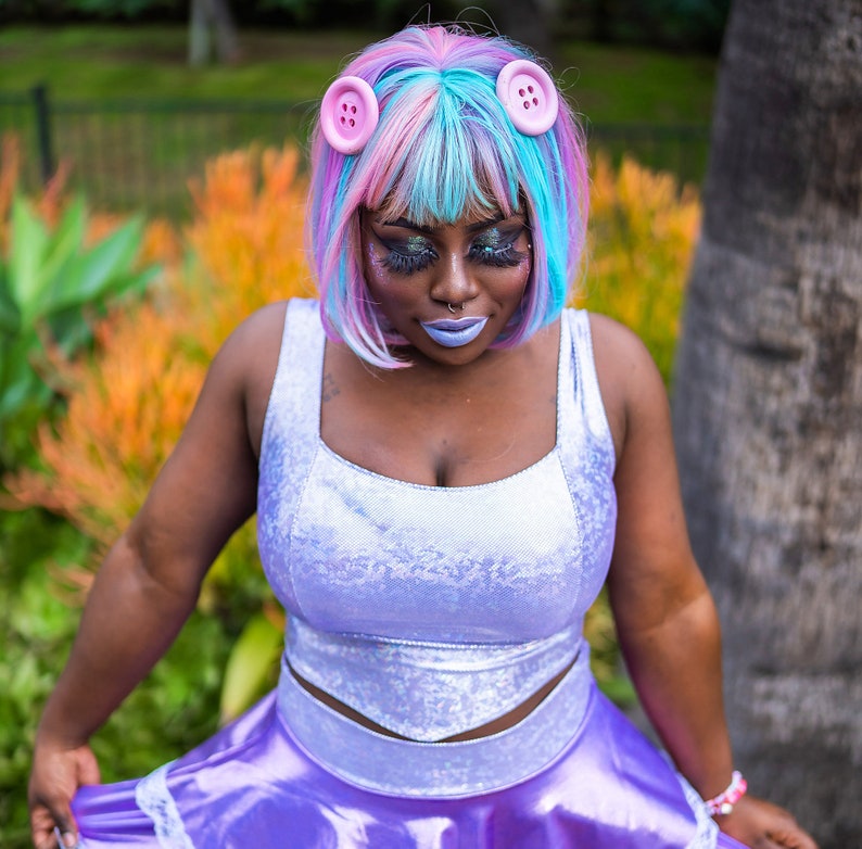 May include: A woman with pink and blue hair wearing a light purple, sparkly, sleeveless dress with a white bodice and a ruffled skirt. She is wearing pink buttons in her hair.