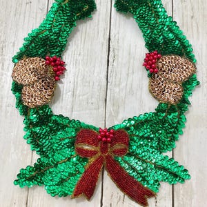 Può includere: Un festoso design di ghirlanda natalizia, con una base di paillettes verdi. Presenta abbellimenti dorati, grappoli di bacche rosse e un fiocco rosso perlato. Perfetto per lavori artigianali e decorazioni stagionali.