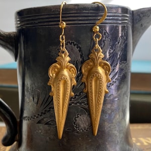 Könnte beinhalten: Ein Paar goldfarbene Ohrhänger. Jeder Ohrring hat ein verziertes, symmetrisches Design mit einem dekorativen Oberteil und einem langen, spitz zulaufenden Boden. Die Ohrringe sind an goldfarbenen Ohrhaken befestigt.