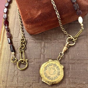 Peut inclure: Collier doré ancien avec un médaillon rond orné de motifs floraux et d'un écusson. Le collier comprend une chaîne, des perles rouge foncé et une perle ovale blanche. Le médaillon est fixé à un fermoir décoratif.