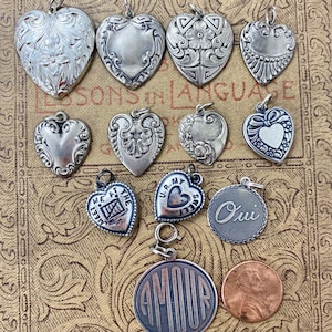 Puede incluir: Una colección de colgantes plateados en forma de corazón con varios diseños, incluyendo patrones florales y detalles grabados. Algunos colgantes tienen texto, como "Oui" y "Amour". Se incluye una moneda de un centavo como referencia de tamaño.