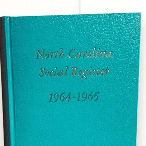 May include: A stack of vintage books, including a teal hardcover book titled "North Carolina Social Register 1964-1965." Other books in the stack have blue, green, and red covers. The books are arranged on a white background.