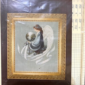 May include: Framed cross-stitch artwork featuring an angel holding a globe. The angel has brown hair, white wings, and wears a blue and gray gown. The artwork is in a gold-colored ornate frame, set against a brown background.
