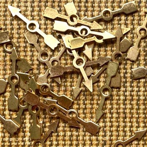 May include: A pile of gold-colored metal arrow-shaped clock hands with a small hole in the center of each hand.