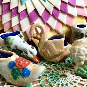 Pode incluir: Cinco almofadas de alfinetes de cerâmica em forma de cisne, pato, sapato, coruja e urso. As almofadas de alfinetes são decoradas com padrões florais e estão sobre uma renda de crochê.