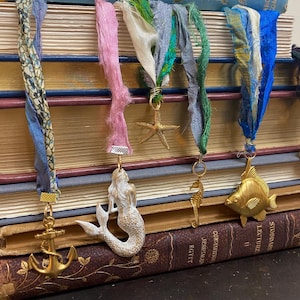 May include: A collection of handmade bookmarks with nautical-themed charms. Each bookmark features a colorful ribbon and a gold-toned charm, including an anchor, mermaid, starfish, seahorse, and fish. The bookmarks are displayed on a stack of vintage books.
