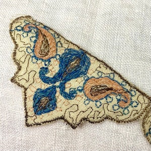 Puede incluir: Un parche de tela beige con bordado de paisley azul y marrón. El parche tiene un borde festoneado.