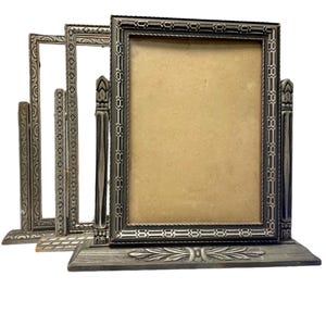 May include: A collection of antique silver-toned picture frames. The frames have ornate detailing, including rope-like borders and decorative columns. The frames are arranged in a row, with the largest frame in the center. The frames are on a white background.