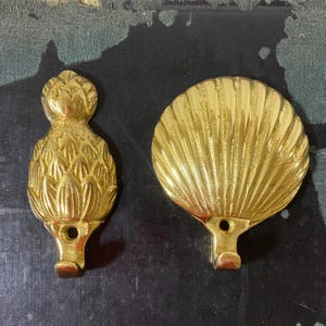 May include: Two brass decorative hooks. One hook is shaped like a pineapple, the other is a seashell. Both hooks have a loop for hanging and a screw hole for mounting. The hooks are gold in colour.