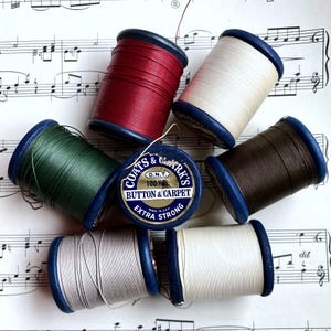 Pode incluir: Uma coleção de bobinas de linha coloridas, incluindo vermelho, verde, creme e castanho, com bordas azuis. Uma bobina central diz "COATS & CLARK'S BUTTON & CARPET EXTRA STRONG". As bobinas estão dispostas em uma partitura musical.