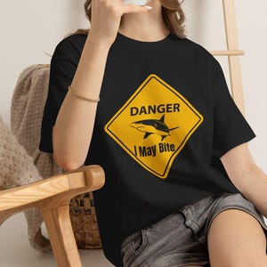 以下が含まれることがあります： 黒の半袖Tシャツ。道路標識を模した黄色のひし形のグラフィックがプリントされています。グラフィックには黒いサメのシルエットが描かれており、その上に「DANGER」、下に「I May Bite」という文字が太い黒字で記されています。
