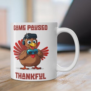 May include: White ceramic mug featuring a cartoon turkey wearing a VR headset and holding a game controller. The mug has the text "GAME PAUSED" above the turkey and "THANKFUL" below. The turkey is brown with red feathers.