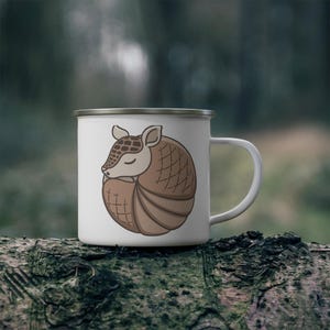 May include: White enamel camping mug with a brown and tan cartoon armadillo curled up sleeping. The mug has a silver rim and handle.
