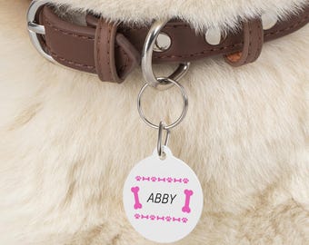 Op de afbeelding: Ronde witte hondenpenning met roze bot- en pootprintontwerpen. De penning heeft de tekst "Abby" in roze letters.