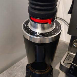 Op de afbeelding: Een zwarte koffiemolen met een houten bovenkant en een rode verstelhendel. De molen heeft een zwarte rubberen balg en een zilveren wijzerplaat met cijfers en tekst. De molen is ontworpen voor espresso.