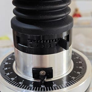 Op de afbeelding: Close-up van een koffiemolen met een zwarte behuizing en een zilverkleurige basis. De molen heeft een wijzerplaat met cijfers en een hendel om de maalgrootte aan te passen. De bovenkant is van hout en zwart rubber.