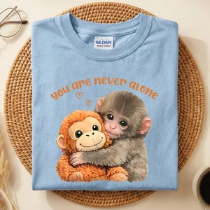 Puede incluir: Camiseta azul claro con un gráfico de dos monos abrazándose y el texto "you are never alone" en naranja. La camiseta es de algodón grueso y está doblada sobre una esterilla tejida.