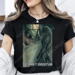Puede incluir: Camiseta negra con una ilustración de fantasía de un hombre lobo y una persona tocándose las manos. La imagen incluye una ventana y el texto "THEY DON'T UNDERSTAND". La obra de arte es en tonos grises y verdes.