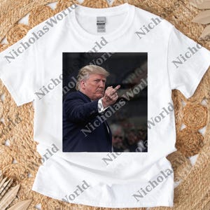 May include: White t-shirt featuring a centered image of a man in a dark suit pointing his middle finger. The image is a close-up of the man's face and hand. The shirt has a crew neck and short sleeves. The brand name "GILDAN" is visible on the shirt's tag.