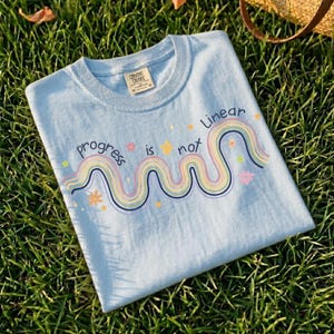 May include: A light blue t-shirt with the words "progress is not linear" in a whimsical font. The design includes a colorful, wavy rainbow and small flower accents. The shirt is made of a soft material and is laying on a bed of green grass.