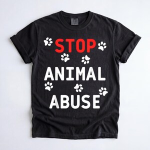 Könnte beinhalten: Schwarzes T-Shirt mit den Worten "STOP ANIMAL ABUSE" in weißen Blockbuchstaben und rotem Text. Weiße Pfotenabdrücke sind um den Text herum verstreut. Das Shirt hat hochgekrempelte Ärmel.