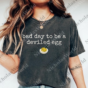 Può includere: T-shirt grigio scuro con la scritta bianca "bad day to be a deviled egg". Sotto c'è un'illustrazione di un uovo farcito. La maglietta è uno stile casual unisex.