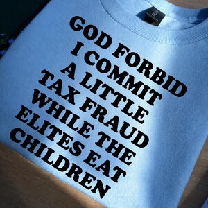 May include: A light blue crewneck sweatshirt featuring bold black text that reads: GOD FORBID I COMMIT A LITTLE TAX FRAUD WHILE THE ELITES EAT CHILDREN. The garment has a ribbed collar and a standard fit, showcasing a clear, high-contrast graphic print.