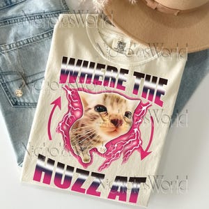 Op de afbeelding: Crèmekleurig T-shirt met een afbeelding van een katje en de tekst "WHERE THE HUZZ AT" in een roze en zwarte gradiënt. Het ontwerp bevat roze bliksemvormen en pijlen. Het T-shirt ligt op een blauwe jeans.