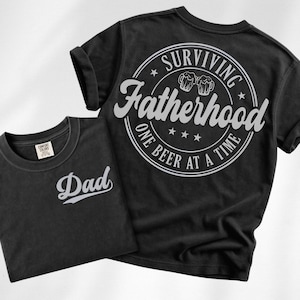 May include: Two black t-shirts. One shirt has the word "Dad" in white script. The other shirt has a white circular design with the words "Surviving Fatherhood One Beer At A Time" and beer mugs.