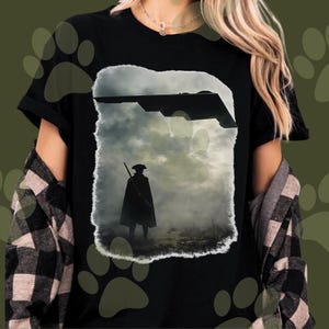 Könnte beinhalten: Schwarzes T-Shirt mit einer Grafik einer Silhouette einer Person mit Dreispitz und Umhang, die ein Gewehr hält, unter einem Tarnkappenbomber vor bewölktem Himmel. Das Design hat einen Used-Look.