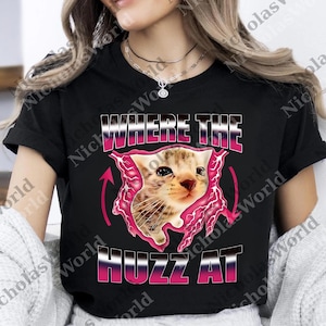 May include: Black t-shirt featuring a kitten graphic with the text "WHERE THE HUZZ AT" in pink and white. The kitten appears to be breaking through a pink, jagged frame. A fun and playful design.