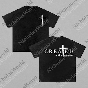 May include: Two black t-shirts with white cross designs. One shirt has a cross on the front, the other has a cross and the words "CREATED with a purpose" on the back. The shirts have a vintage, washed look.