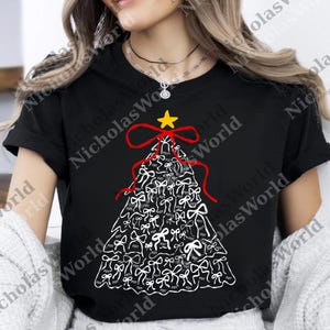 Può includere: T-shirt nero con un design a forma di albero di Natale composto da fiocchi bianchi, un nastro rosso e una stella gialla. I fiocchi hanno le lettere "FOT" stampate sopra. Una maglietta grafica festiva per le festività.