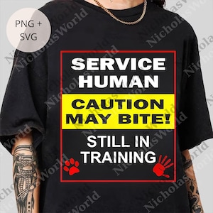 Puede incluir: Camiseta negra con el texto "SERVICE HUMAN CAUTION MAY BITE! STILL IN TRAINING." El diseño incluye una huella de pata roja y una huella de mano roja. Es una prenda informal.
