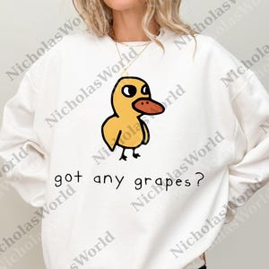 Op de afbeelding: Witte sweatshirt met een cartoon eend. De eend heeft een gele body, zwarte ogen en een oranje snavel. De tekst "got any grapes?" staat eronder in het zwart. Een casual, comfortabel kledingstuk.