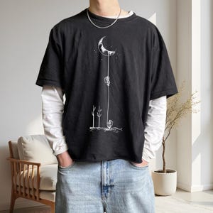 May include: A black short-sleeve t-shirt layered over a white long-sleeve shirt. The black t-shirt features a white line art graphic of an astronaut standing on the ground, connected by a long rope to another astronaut hanging from a crescent moon.