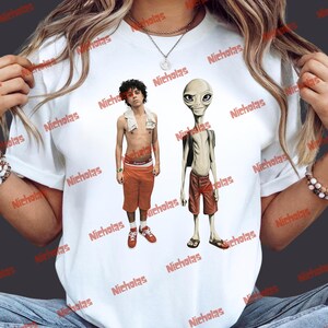 May include: White t-shirt featuring a graphic of two figures: a person in orange shorts and sneakers, and an alien in red shorts and sandals. The shirt has the word "Nicholas" printed repeatedly.