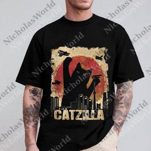 May include: Black t-shirt featuring a silhouette of a cat in a Godzilla-like pose, with a red sun and city skyline backdrop. The text "CATZILLA" is displayed in gold. The design includes airplanes.
