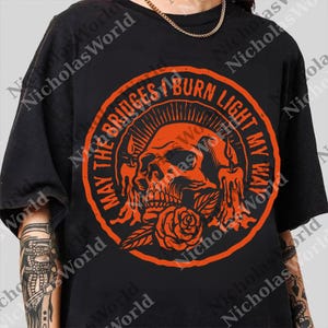 May include: Black t-shirt with an orange graphic. The graphic features a skull, rose, and candles within a circular design. Text around the circle reads "MAY THE BRIDGES I BURN LIGHT MY WAY."