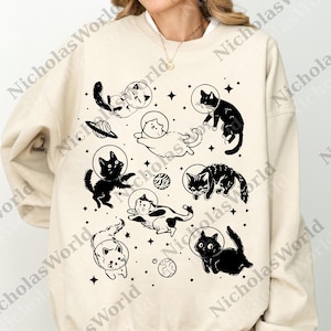 May include: Beige sweatshirt featuring a black and white illustration of cats in space helmets, planets, and stars. The design includes various cat poses, creating a whimsical and playful aesthetic. The sweatshirt is a comfortable, casual garment.