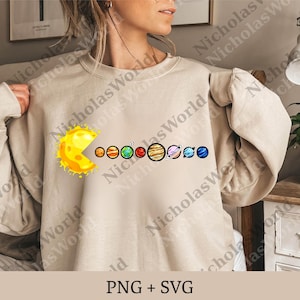 May include: Beige sweatshirt featuring a yellow sun graphic eating the planets of the solar system. The planets are in a row, each with its unique colors and features. The text "PNG + SVG" is at the bottom.