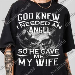 以下が含まれることがあります： 「GOD KNEW I NEEDED AN ANGEL SO HE GAVE ME MY WIFE」の白文字がプリントされた黒いTシャツ。グラフィックは、お互いを握り合う2つの手を示しています。上部には金のチェーンが見えます。