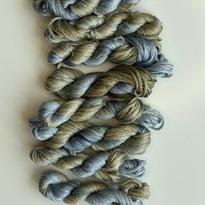 May include: A collection of seven twisted skeins of embroidery thread, each featuring a variegated blend of muted sage green and soft periwinkle blue fibers. The skeins are arranged in a vertical stack, showcasing the twisted texture of the thread.