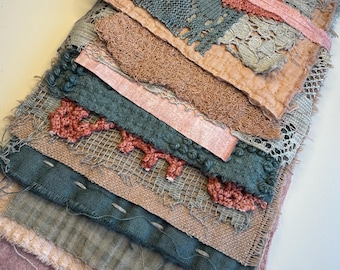 May include: A layered textile collage featuring various fabric scraps in shades of dusty blue, muted pink, and sage green. The composition includes pieces of lace, linen, and textured woven materials, all held together by a thin, light pink ribbon tie.