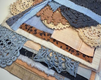 May include: A stack of assorted fabric scraps and crochet doilies in shades of beige, brown, blue, and black, tied together with a light blue fabric ribbon. The collection includes various textures, floral prints, and intricate lace patterns.