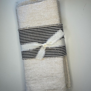 May include: A stack of folded, off-white, textured fabric napkins secured with a band of black and white striped fabric. The band is tied in the center with a frayed, off-white fabric ribbon, creating a simple, layered presentation of textiles.
